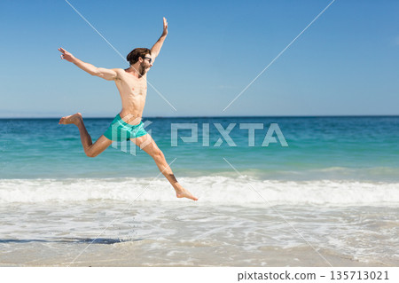 Man wearing turquoise swim shorts and dark sunglasses leaping above surf on sandy beach, copy space Man wearing turquoise swim shorts and dark sunglasses leaping above surf on sandy beach, copy space 135713021