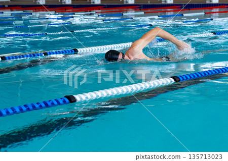 Male swimmer performing front crawl at aquatic center pool with swim cap, goggles and lane ropes Male swimmer performing front crawl at aquatic center pool with swim cap, goggles and lane ropes 135713023