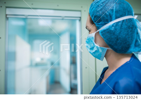 Female nurse wearing scrubs standing by glass door gazing into OR in surgical wing, copy space 135713024
