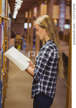 Teenage female student reading open book between bookshelves in library aisle under warm lighting Teenage female student reading open book between bookshelves in library aisle under warm lighting 135713036