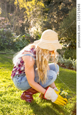 Female gardener kneeling in backyard garden holding yellow blooms wearing garden gloves copy space Female gardener kneeling in backyard garden holding yellow blooms wearing garden gloves copy space 135713048