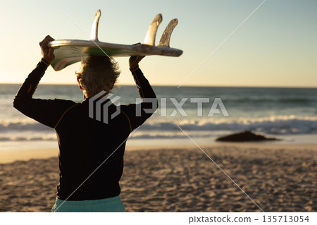 Senior African American woman in wetsuit carrying surfboard overhead on beach at sunset with rock 135713054