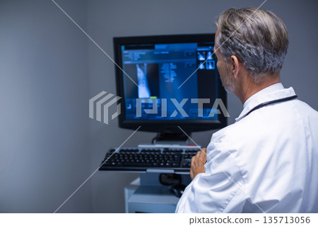 Male doctor reviewing diagnostic scans on monitor at workstation with stethoscope, copy space 135713056