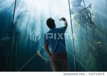 Man in his thirties holding smartphone capturing fish and kelp through aquarium glass at exhibit 135713078