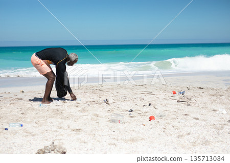 Senior African American man bending over on sandy beach holding trash bag picking bottles Senior African American man bending over on sandy beach holding trash bag picking bottles 135713084