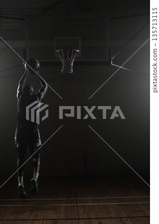 Basketball flying toward hoop in dimly lit gymnasium, spotlights casting dramatic shadows 135713115