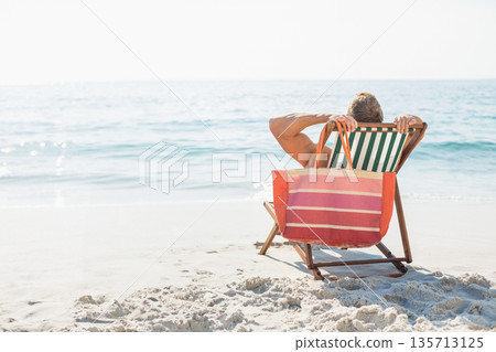 Man relaxing on striped deck chair on sunlit sandy ocean shoreline holding tote bag, copy space 135713125