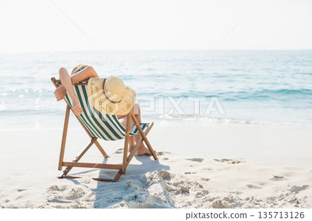 Woman wearing swimsuit sitting on striped beach chair on sandy shoreline holding straw sun hat 135713126