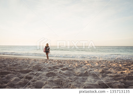 Woman walking along sandy beach at water's edge while holding red surfboard under arm Woman walking along sandy beach at water's edge while holding red surfboard under arm 135713135