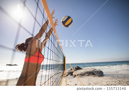 Female athlete in red sportswear reaching to block volleyball over net on beach volleyball court 135713148
