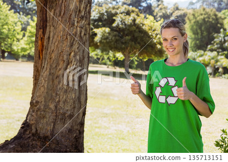 Adult woman standing beside large tree trunk in park, wearing green recycling Tshirt and smiling 135713151