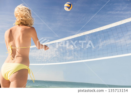 Woman wearing yellow bikini reaching for yellow-and-white volleyball at net on beach, copy space Woman wearing yellow bikini reaching for yellow-and-white volleyball at net on beach, copy space 135713154