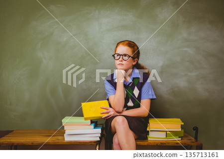 Girl student sitting on bench in classroom holding yellow book amid book stacks and chalkboard Girl student sitting on bench in classroom holding yellow book amid book stacks and chalkboard 135713161