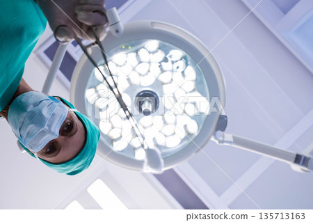Female surgeon wearing mask leaning in operating room holding forceps under LED light, copy space Female surgeon wearing mask leaning in operating room holding forceps under LED light, copy space 135713163