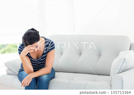 Mature woman leaning forward on gray sofa in living room under white blinds wearing T-shirt, jeans 135713164