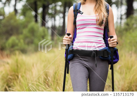 Woman trekking through meadow at pine forest edge holding trekking poles and wearing blue backpack 135713170