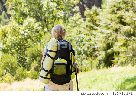 Senior woman hiking along sunlit forest trail carrying large backpack and gripping trekking pole Senior woman hiking along sunlit forest trail carrying large backpack and gripping trekking pole 135713171