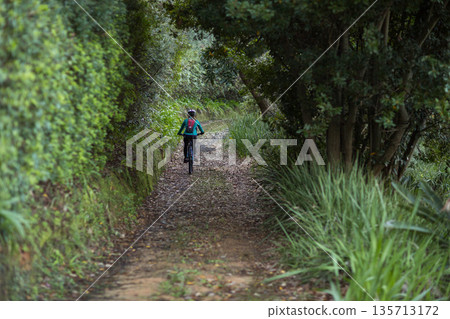 Woman cycling on mountain bike wearing helmet along dirt path bordered by stone edging in forest Woman cycling on mountain bike wearing helmet along dirt path bordered by stone edging in forest 135713172