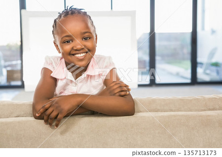 African American girl leaning over sofa back smiling at camera in living room through window panels 135713173