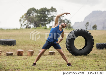 Male fitness enthusiast stretching in open field beside stumps and tractor tires with running shoes Male fitness enthusiast stretching in open field beside stumps and tractor tires with running shoes 135713180