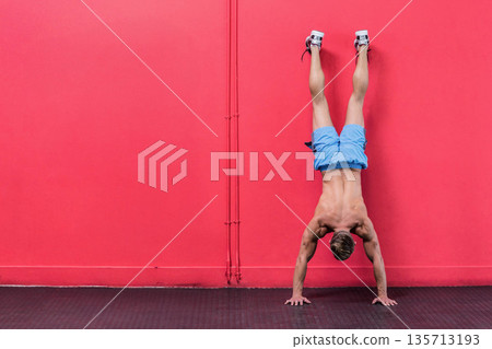 Male athlete balancing upside-down against red wall on rubber mat wearing blue shorts, copy space 135713193