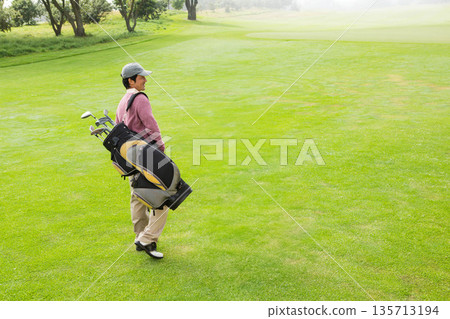 Mid adult Japanese man in golf attire walking across fairway carrying bag with clubs, copy space Mid adult Japanese man in golf attire walking across fairway carrying bag with clubs, copy space 135713194