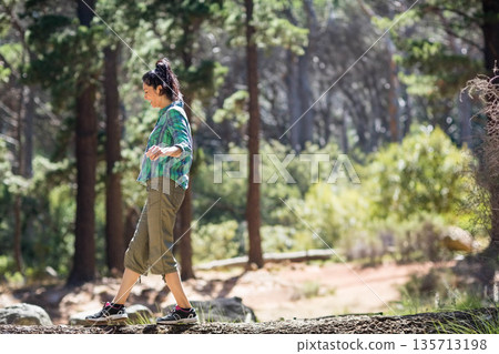 Woman balancing on fallen log through sunlit pine forest clearing with dirt trail and rocks Woman balancing on fallen log through sunlit pine forest clearing with dirt trail and rocks 135713198