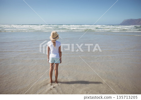 Girl standing in wet sand at shoreline wearing straw sun hat and gazing at ocean waves Girl standing in wet sand at shoreline wearing straw sun hat and gazing at ocean waves 135713205