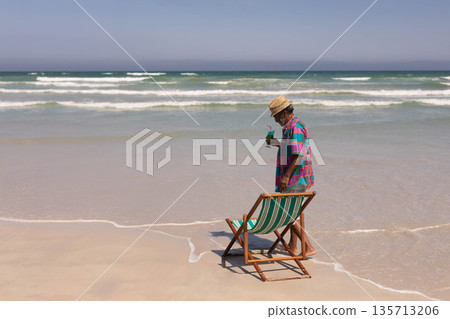 Senior man holding cold drink wearing straw hat sunglasses at shore by deck chair, copy space Senior man holding cold drink wearing straw hat sunglasses at shore by deck chair, copy space 135713206