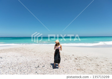 Woman standing on pebbly beach wearing sun hat and purple swimsuit while wrapping black sarong Woman standing on pebbly beach wearing sun hat and purple swimsuit while wrapping black sarong 135713209