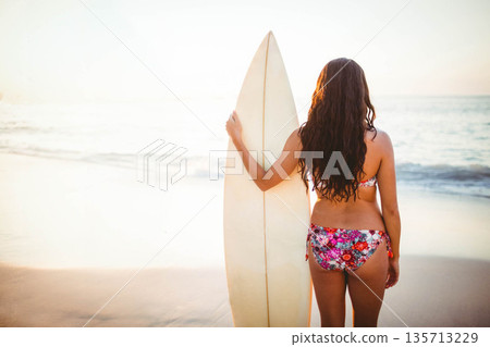 Female surfer wearing floral bikini standing on shore at sunset holding cream surfboard, copy space Female surfer wearing floral bikini standing on shore at sunset holding cream surfboard, copy space 135713229