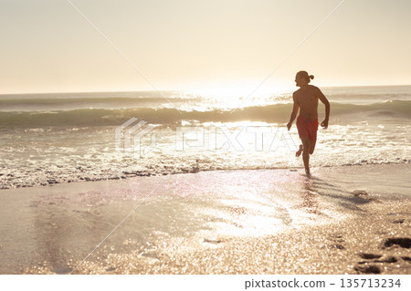 Athletic male sprinting along sandy beach shoreline under golden sunlight reflections, copy space 135713234
