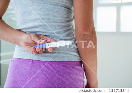 Woman standing facing away holding pregnancy test behind back in softly lit private room Woman standing facing away holding pregnancy test behind back in softly lit private room 135713244