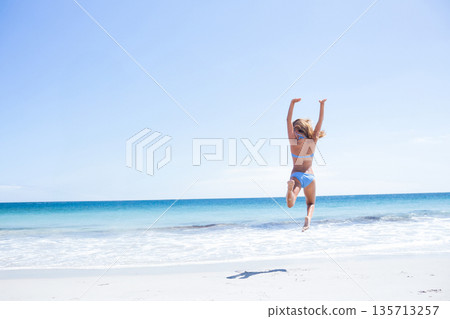 Female beachgoer jumping mid-air on sandy beach under clear blue sky with turquoise ocean waves 135713257