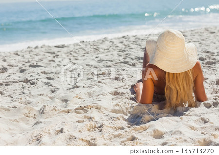 Woman lying on white sand wearing bikini, straw hat gazing toward turquoise waves, copy space Woman lying on white sand wearing bikini, straw hat gazing toward turquoise waves, copy space 135713270