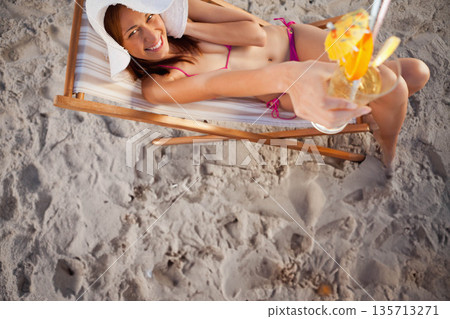 Woman reclining on beach chair wearing pink bikini and hat extending cocktail glass with umbrella Woman reclining on beach chair wearing pink bikini and hat extending cocktail glass with umbrella 135713271