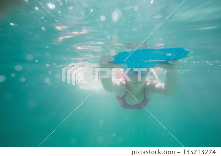 Elementary school girl swimming underwater in pool gripping blue foam kickboard and wearing goggles Elementary school girl swimming underwater in pool gripping blue foam kickboard and wearing goggles 135713274