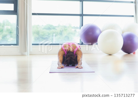 Woman in pink sportswear practicing yoga on purple mat in studio with large windows, exercise balls 135713277
