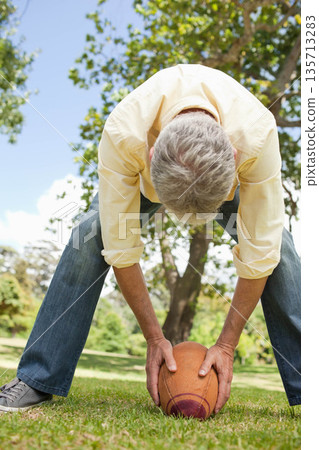 Male bending over placing American football on park grass under blue sky wearing pale yellow shirt 135713283