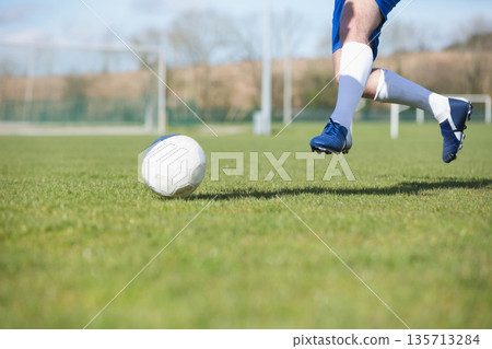 Teen male soccer player wearing blue cleats kicking soccer ball on field near goalpost, copy space 135713284