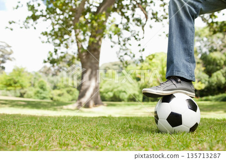Male balancing foot on soccer ball, wearing denim pants, gray sneakers on park lawn, copy space Male balancing foot on soccer ball, wearing denim pants, gray sneakers on park lawn, copy space 135713287