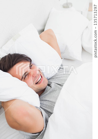 Man wearing gray t-shirt lying on bed and smiling in bedroom with pillows and lamp base 135713297