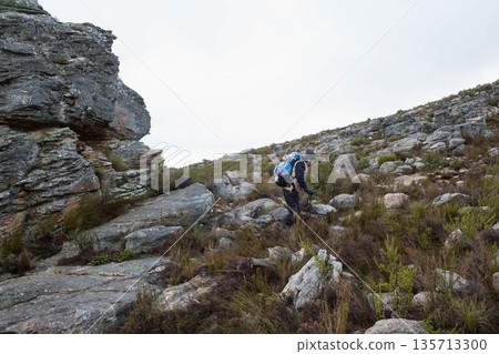 Male hiker wearing beanie climbing rocky slope using trekking pole and backpack amid grey sky Male hiker wearing beanie climbing rocky slope using trekking pole and backpack amid grey sky 135713300