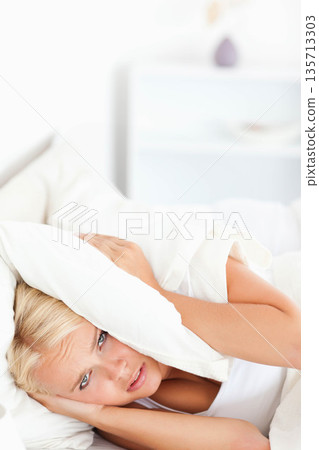 Female wearing white tank top lying on mattress pressing white pillow over ears near bedside vase 135713303