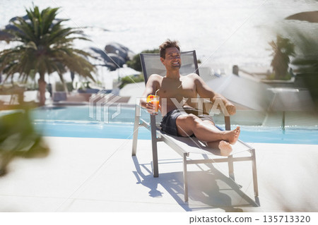 Mid-adult man reclining on metal lounge chair at poolside holding bright orange tropical cocktail 135713320