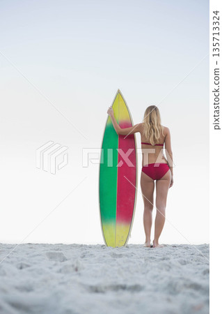 Woman wearing red bikini holding striped surfboard standing on gray sandy beach under pale sky Woman wearing red bikini holding striped surfboard standing on gray sandy beach under pale sky 135713324