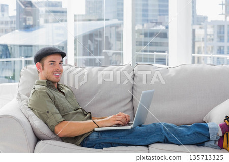 Male reclining on light gray sofa in modern lounge typing on silver laptop, copy space Male reclining on light gray sofa in modern lounge typing on silver laptop, copy space 135713325