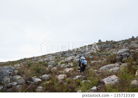 Man in dark jacket hiking uphill across rocky slope carrying blue grey backpack, copy space Man in dark jacket hiking uphill across rocky slope carrying blue grey backpack, copy space 135713330