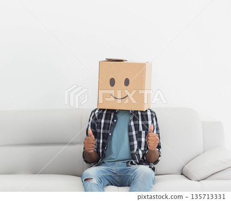 Cardboard box with smiley face resting on light gray sofa against white wall, with white cushion Cardboard box with smiley face resting on light gray sofa against white wall, with white cushion 135713331