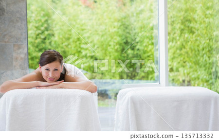Female spa client lying prone on towel-covered massage table near glass wall with green shrubbery 135713332